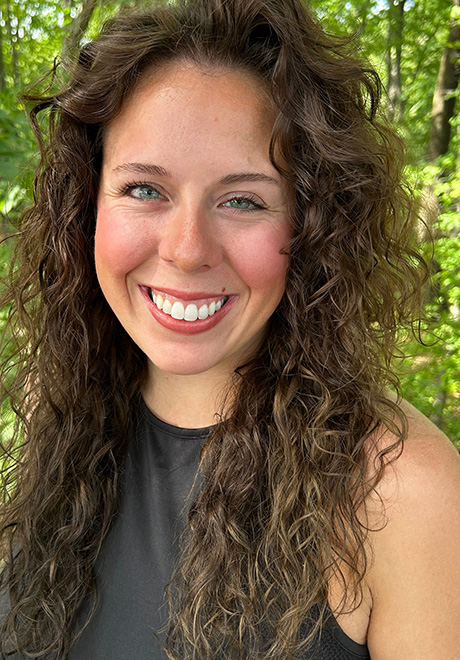 Amber LaBerge headshot: smiling in gray shirt with green leaves in the background.