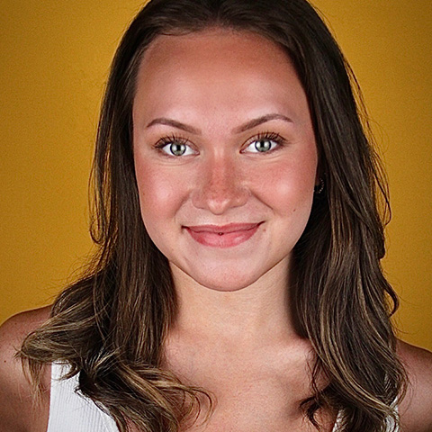 Anna Beyer headshot: grinning in white shirt in front of yellow/tan wall