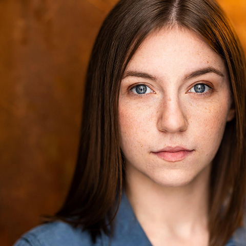 Emma Resek headshot: wearing blue top in front of maroon-brown background