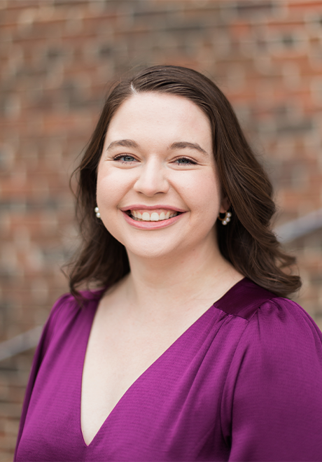 Logan Trotter headshot, wearing a purple v-cut shirt in front of a brick building.