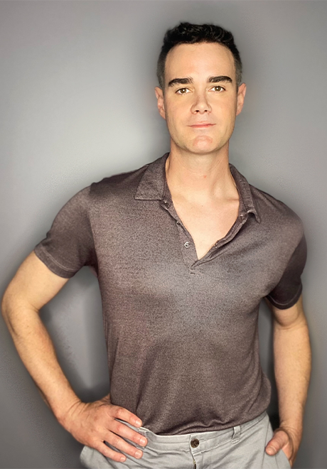 Brett Leigh headshot: holding hand on hip while wearing a metallic-colored shirt in front of a gray wall