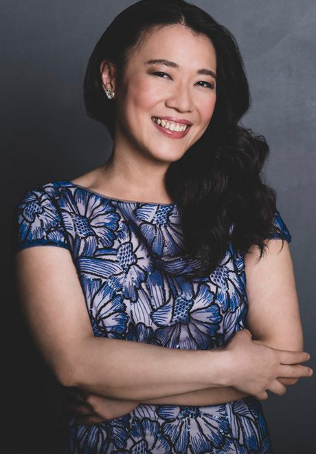 Charlotte Hu headshot. Charlotte is smiling in front of a gray background in a black and white flower-patterned shirt with their arms crossed.