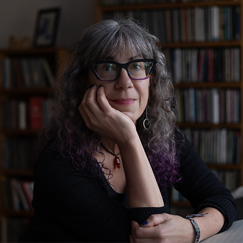 Marti Epstein smiling in front of a book shelf with her hand resting underneath her chin.