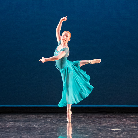 Dancer in blue dress on point with one leg pointed back with arms in the air