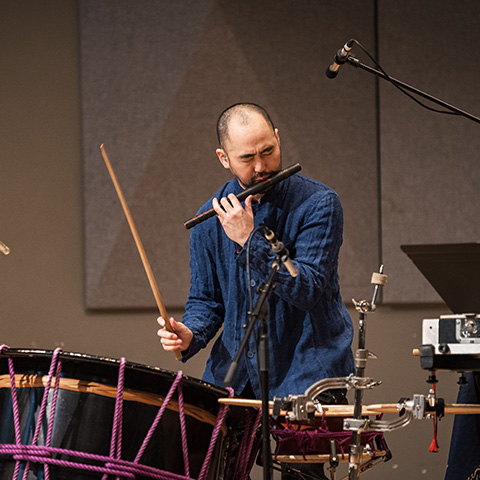 Performer playing a wooden flute with one hand and hitting a drum in the other hand