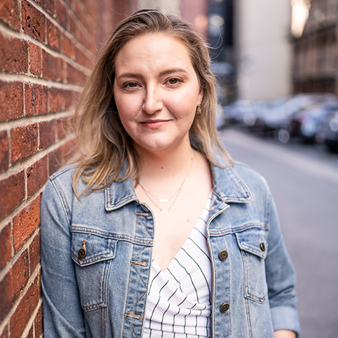 Alison Pugh Headshot: grinning in a jean jacket and white shirt in front of a brick wall.