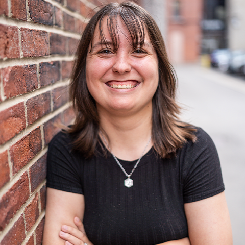 Maddy Poston hedashot: smiling in a black tshirt in front of a brick wall.
