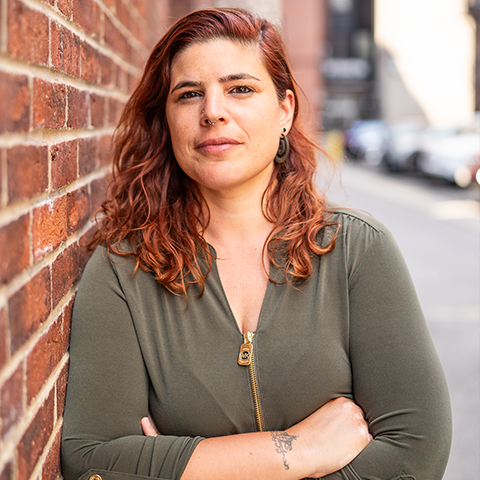 Sara Pagiaro headshot: arms crossed, grinning in an off-green shirt in front of a brick wall.