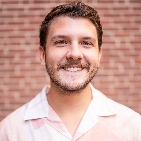 Zachary Sayre headshot: smiling in a light pink button down in front of a brick wall.
