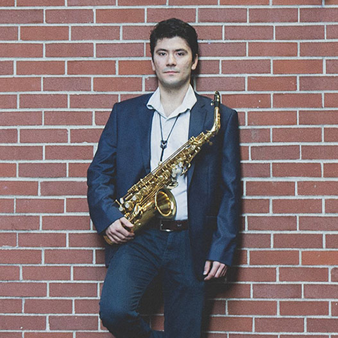Goran Daskalov headshot: holding saxophone and wearing a suit in front of a brick wall.
