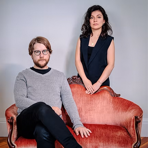 Lamnth, a violin and cimbalom duo, founded in 2023. A woman stands behind a man sitting on a red couch.