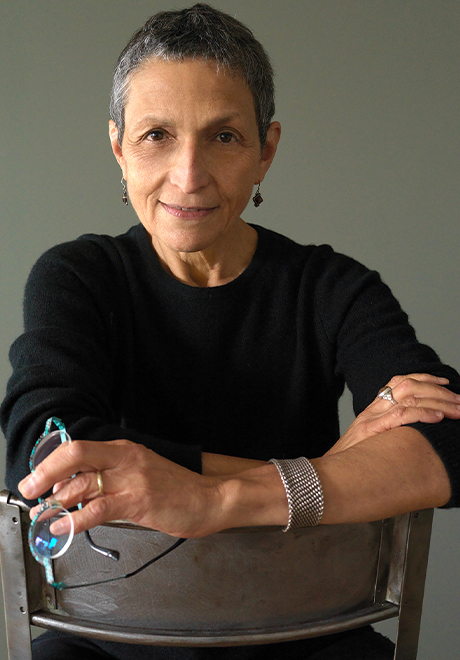 Laura Kaminsky headshot: wearing a black shirt sitting in a chair holding her glasses.