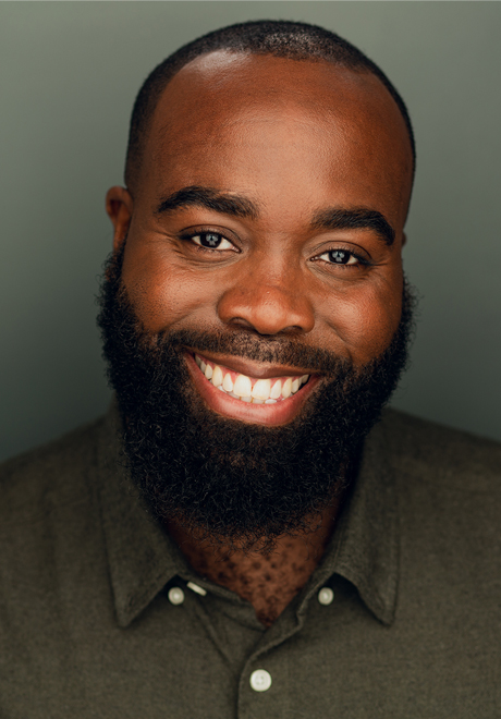 Headshot of Ricardo Coke-Thomas: smiling and wearing a green-gray button down shirt in front of an off-grey background 
