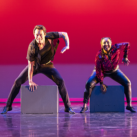 Two dancers on stage, with a hand on a brick that they're standing over. background is red and stage is a reflective purple.