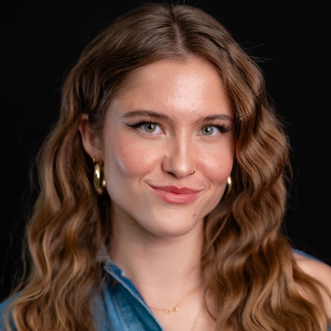 Maisy Davies headshot: smiling in a denim shirt in front of a black background