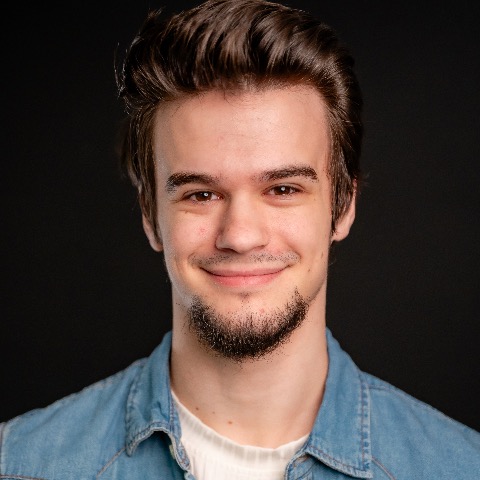 Alex Hutchinon headshot:smiling in a denim shirt and white undershirt in front of a black background.