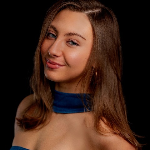 Brook Vass headshot: denim top with a piece of denim wrapped around the neck in front of a black background