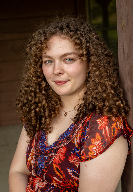 Lily Cooper headshot: smiling wearing a warm-toned shirt in front of a brown background