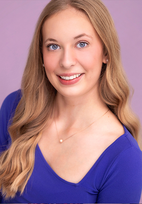 Tahlia Knoshaug headshot:s miling and wearing a blue shirt in front of a pink background
