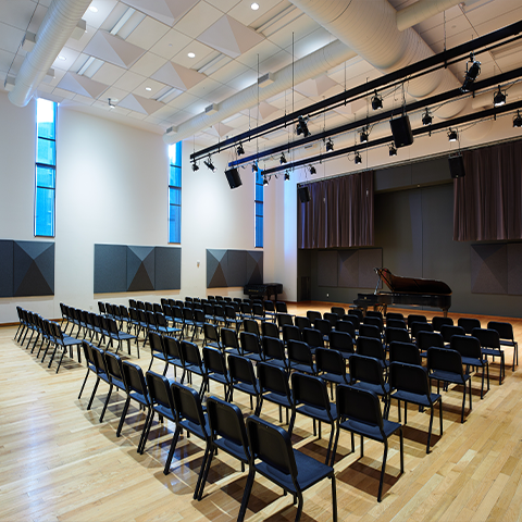 Studio 106, large room with many rows of chairs and a piano at the front of the room. Wooden floors and white walls.