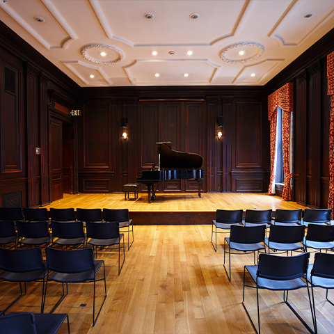 Houston Hall: recital hall with wooden walls and floor, a stage with a piano and chairs in the audience section