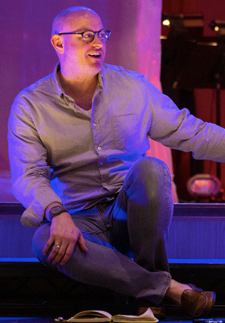 Nathan Troup sitting on stage smiling with his legs half crossed