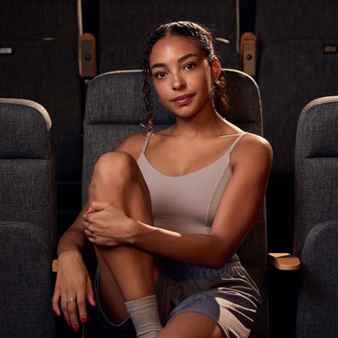 Sonrisa Hubbard headshot: sitting in a theater seat with her left leg pulled into her chest 