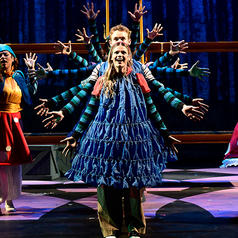 Theater students on stage in blue colorful costumes with their arms out creating a wheel of arms visual