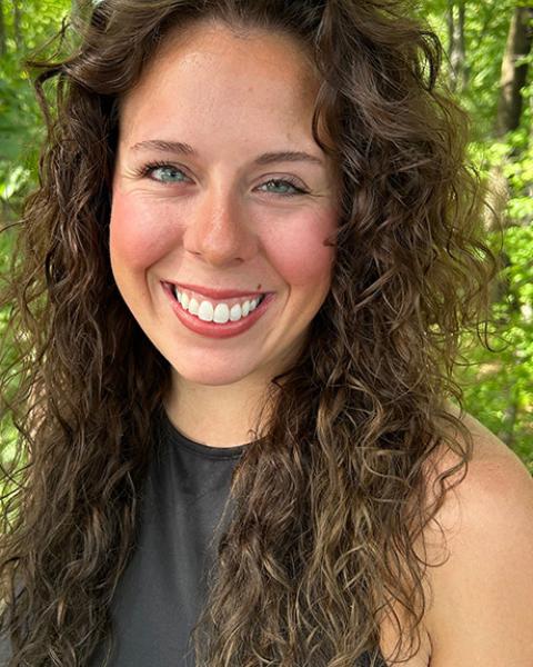 Amber LaBerge headshot: smiling in gray shirt with green leaves in the background.