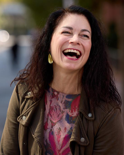 Liz Roncka smiling–mid laugh. She is wearing a colorful (red, blue, purple) shirt with a brown jacket.