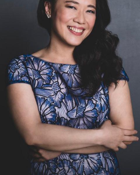 Charlotte Hu headshot. Charlotte is smiling in front of a gray background in a black and white flower-patterned shirt with their arms crossed.