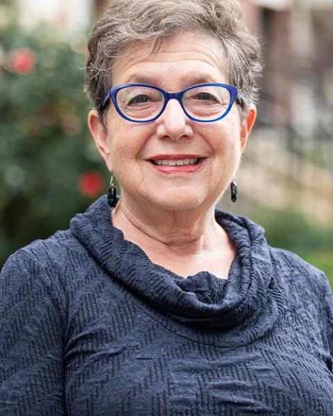 Sara Goldstein headshot: smiling in front of buildings and wearing a blue/gray sweater.
