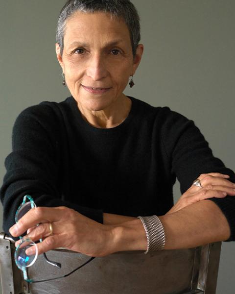Laura Kaminsky headshot: wearing a black shirt sitting in a chair holding her glasses.