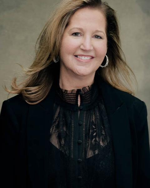 Noel Smith headshot: smiling in all black clothes in front of a gray background.