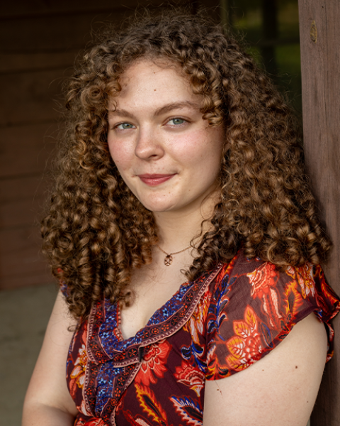Lily Cooper headshot: smiling wearing a warm-toned shirt in front of a brown background