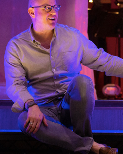 Nathan Troup sitting on stage smiling with his legs half crossed