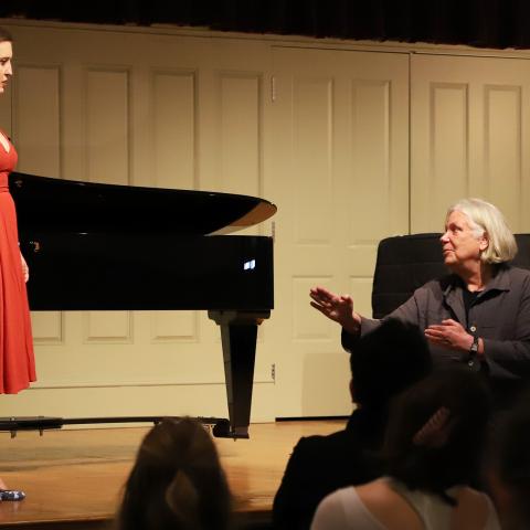 Soprano Sarah DeYong (left, in red dress) and Anne Bogart (right, in black shirt) talking in front of a piano