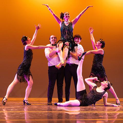 Group of dancers in 1920's style clothing on stage. Two dancers are holding one dancer in the air.