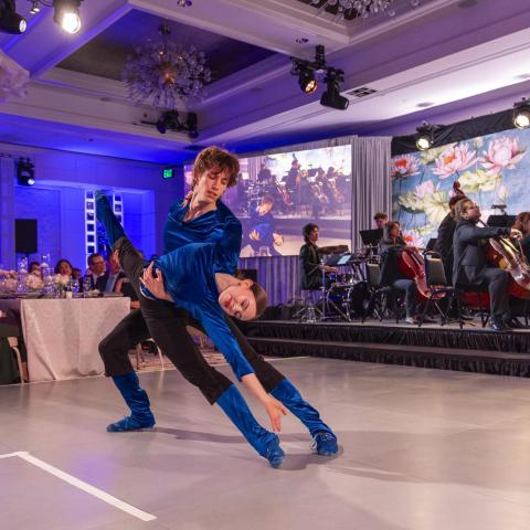 two dance students performing at the Soirée in black and black outfits. One student is holding the other's leg up to the sky.
