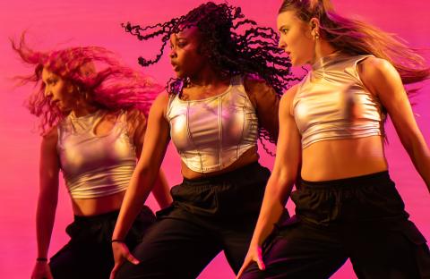 Three dancers in gold tops and black pants whipping their hair to the left in front of a pink background.